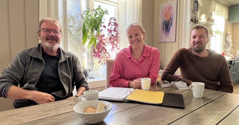 Tre personer ved bord med kaffekopper, perm og notatb&oslash;ker